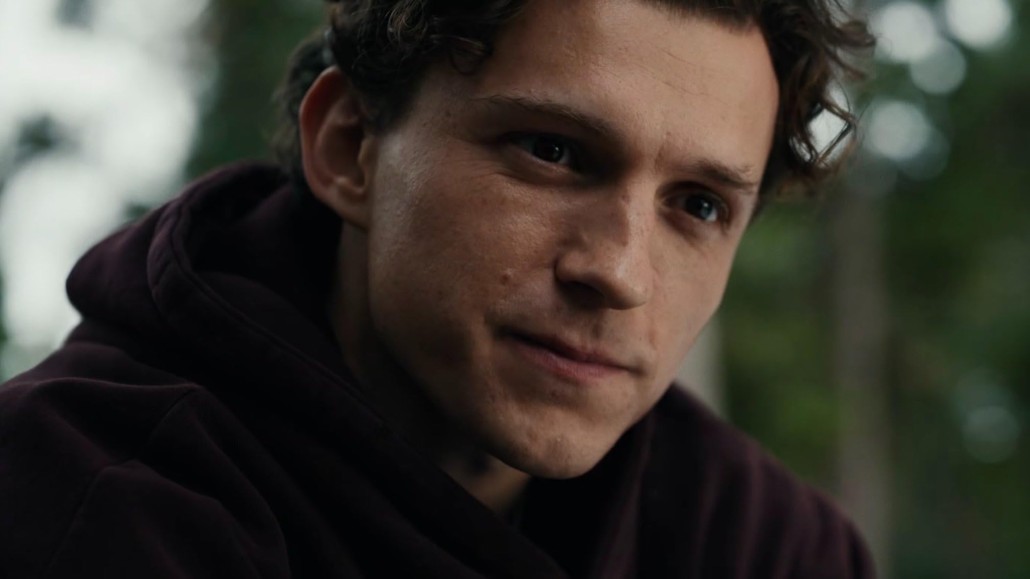 Close-up of a young man with a burgundy hoodie, gazing to the right against a blurred outdoor background.