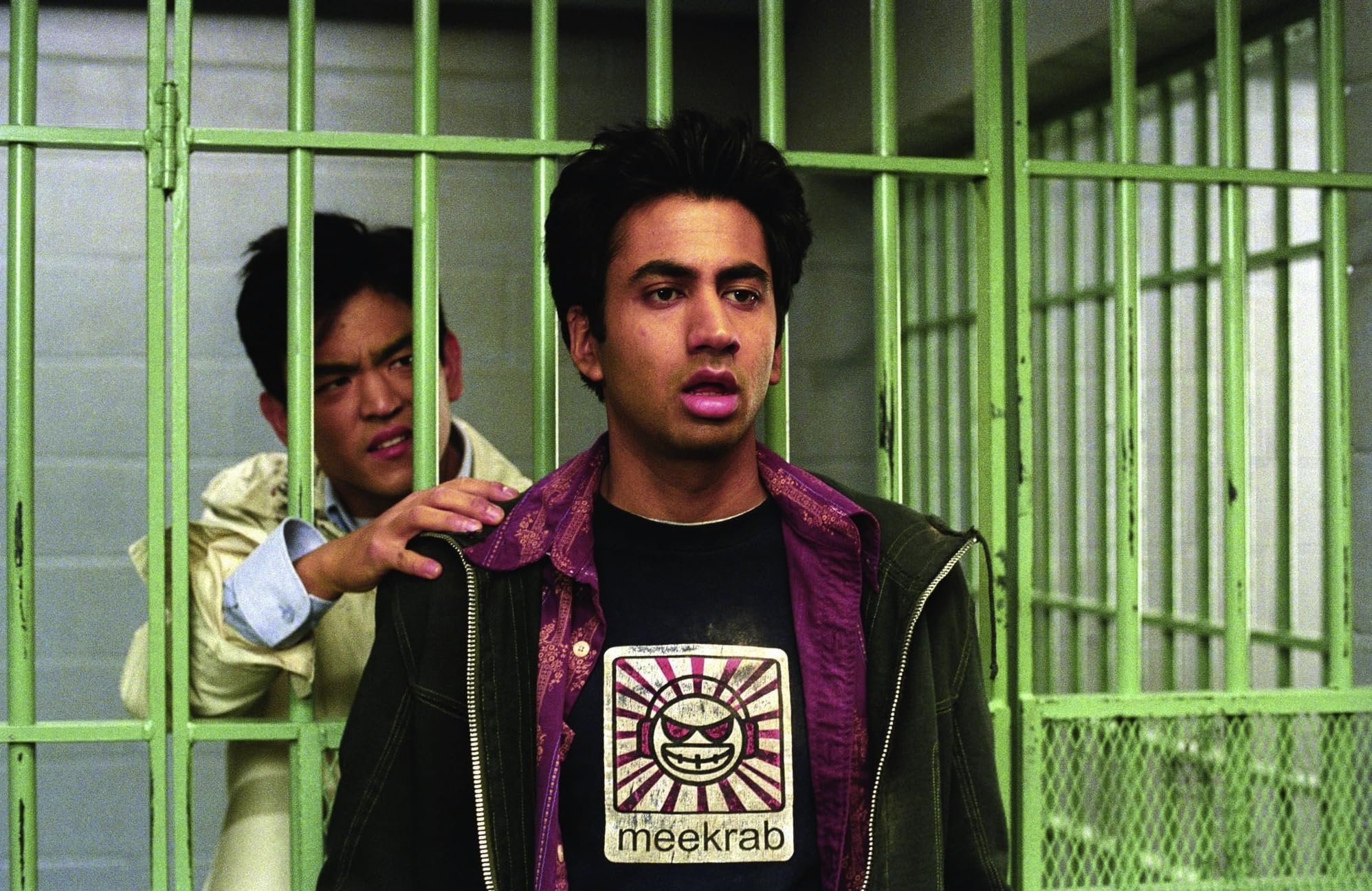 Two men behind a bright green metal barrier; one in the foreground looks surprised while a second man reaches over to touch his shoulder from behind.