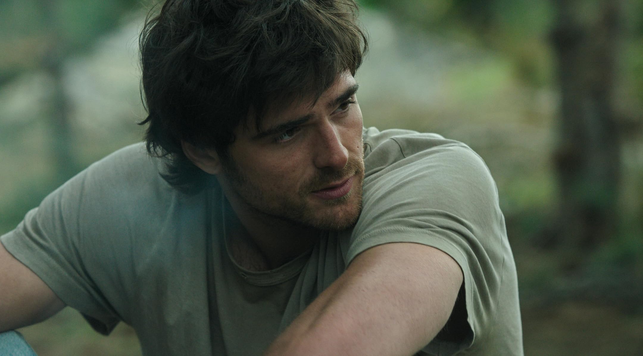 Young man with brown hair and stubble, wearing a light green T-shirt, looking off to the right outdoors.