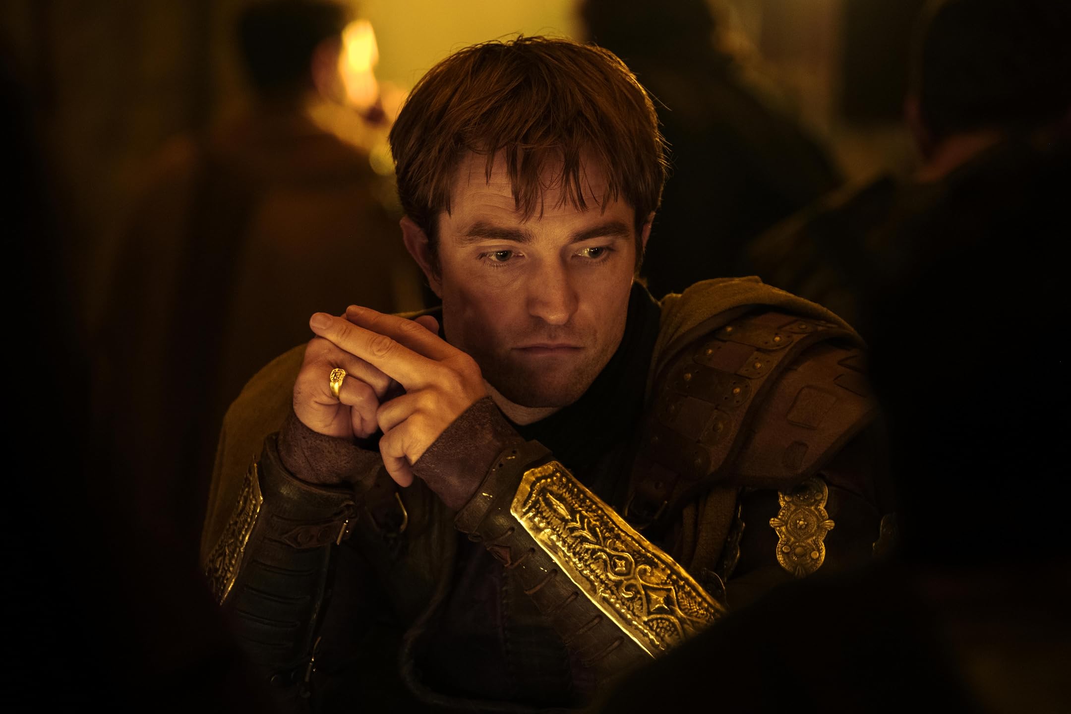 Man wearing ornate medieval armor, clasping his hands in a dim, candlelit setting.