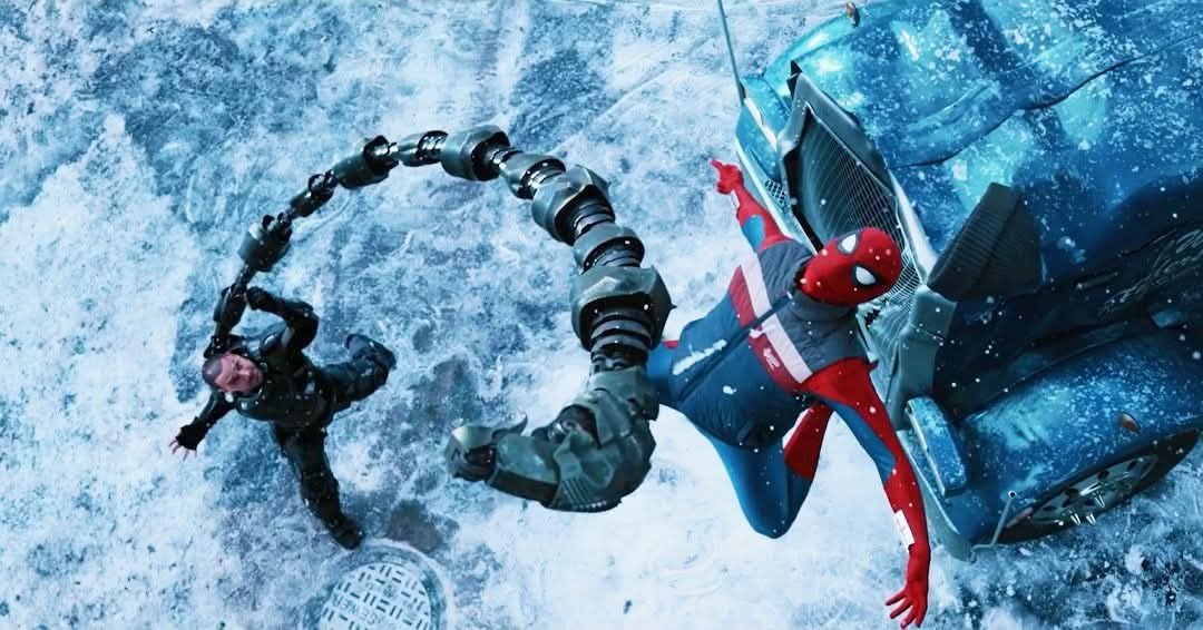 Spider-Man in red and blue suits reaches toward a person on a snowy, icy street beside a damaged car. A jointed, tentacle-like device circles above them as they appear to rescue or assist him.