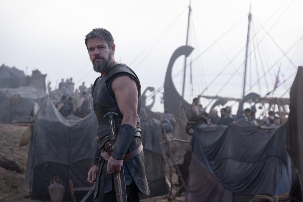Bearded warrior in dark leather armor standing in a dusty camp with tents in the background and ship masts behind him.