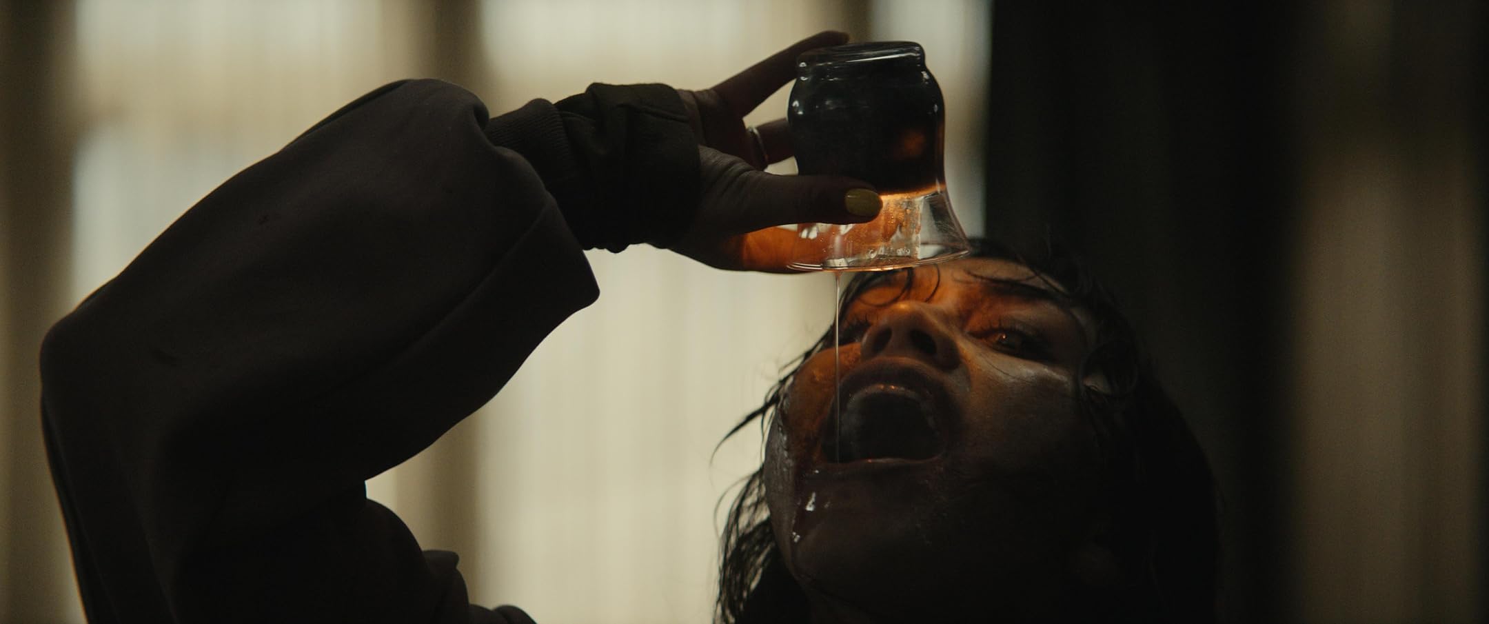 A person tilts a bottle to pour a dark liquid into another person's open mouth in a dim scene.