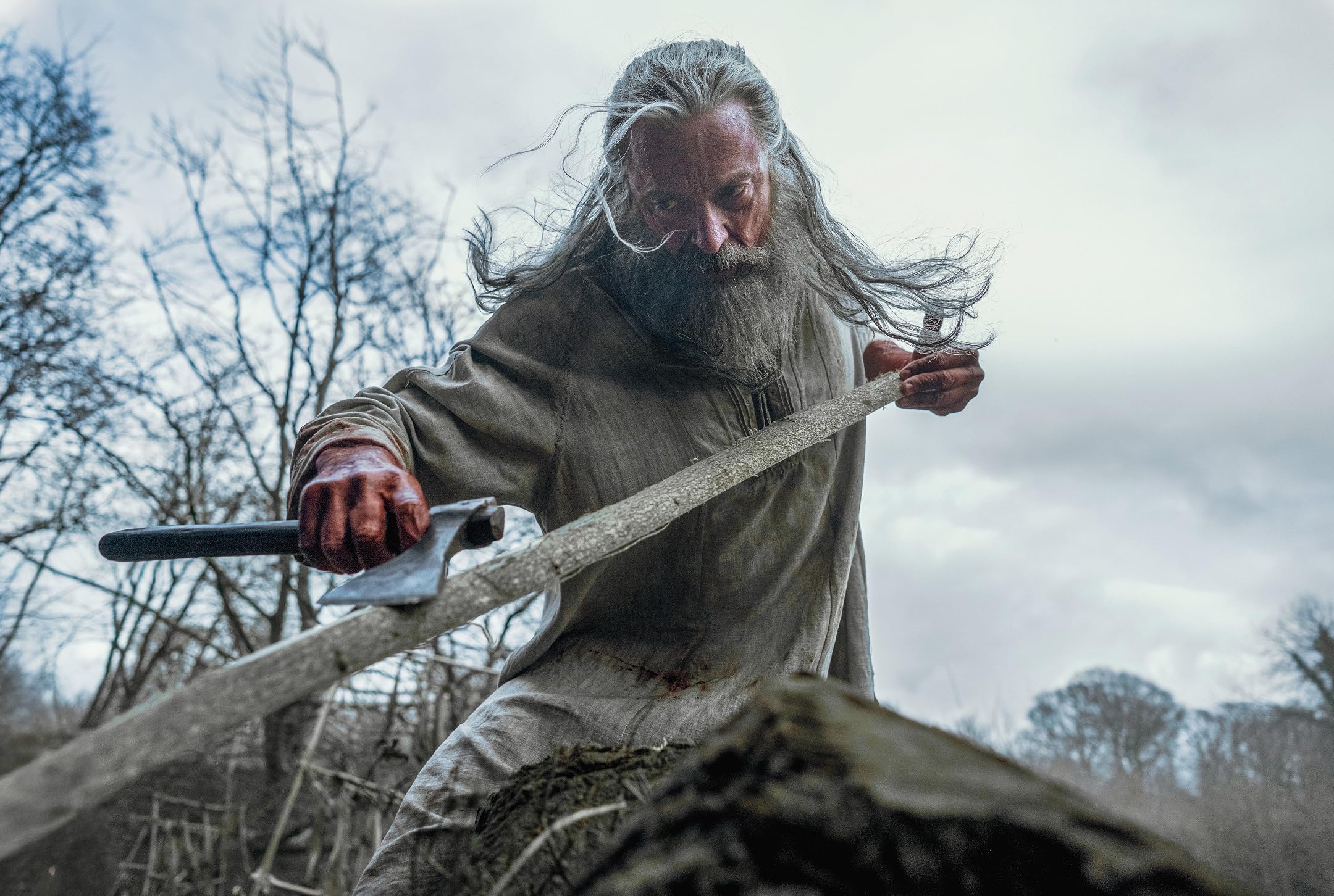 Bearded older man with long gray hair chopping wood outdoors with an axe.
