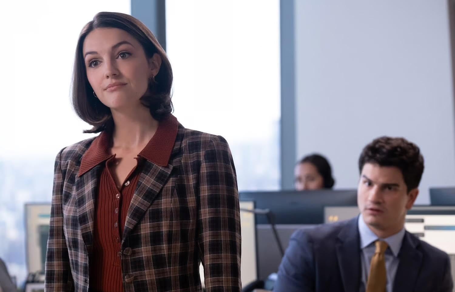 A woman in a plaid blazer stands at the front of an office, looking thoughtful, with two coworkers at desks in the background.