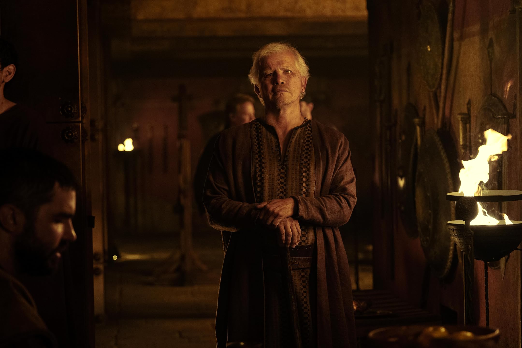 Older man with white hair standing crossed-armed in a dimly lit, medieval-style hall with torches and large stone walls behind him.
