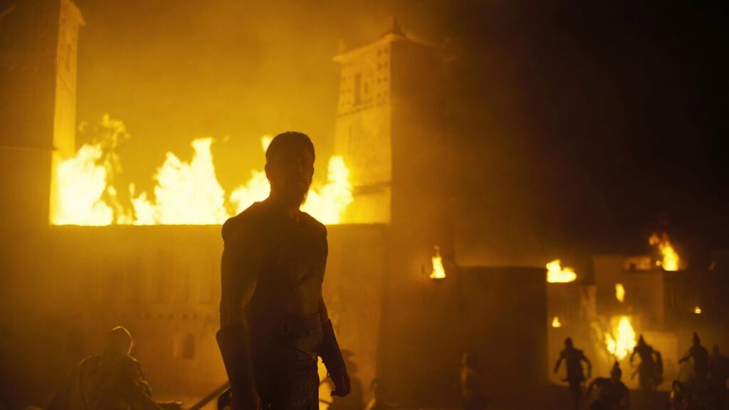 Silhouetted person in a dark costume stands before large flames and burning structures at nightfall festival.