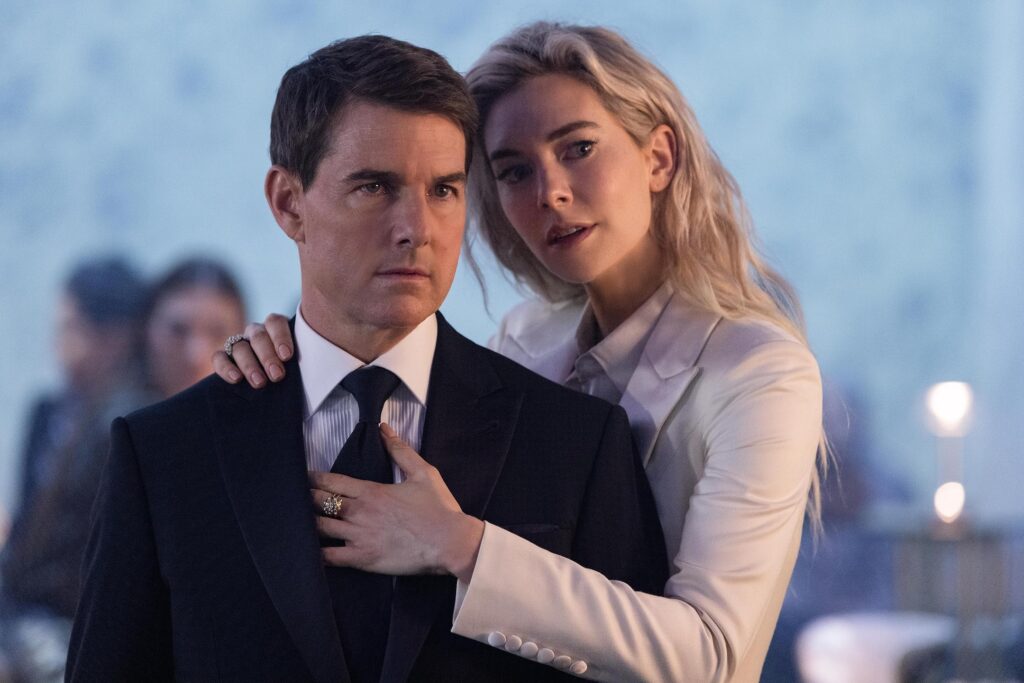 A man in a dark suit and tie stands still while a woman in a light blazer embraces him from behind, resting her hand on his chest with a ring visible; both look thoughtful in a blurred background.