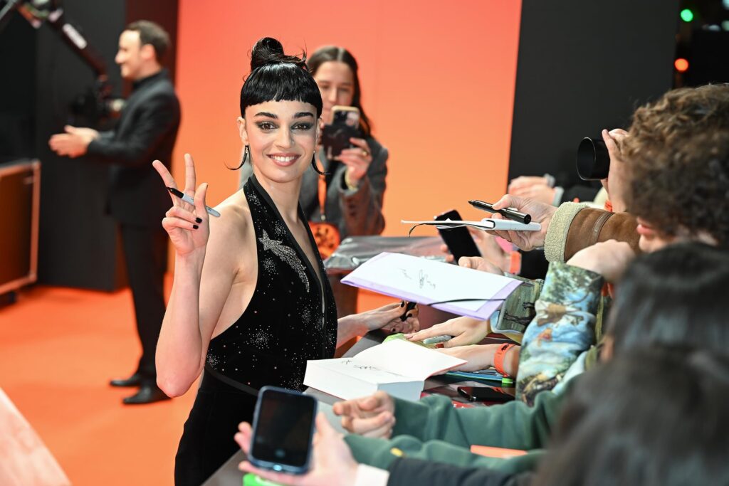 Celebrity woman in a sparkling black dress signs autographs for fans at a fan event against a bright orange backdrop, raising a peace sign with her hand.