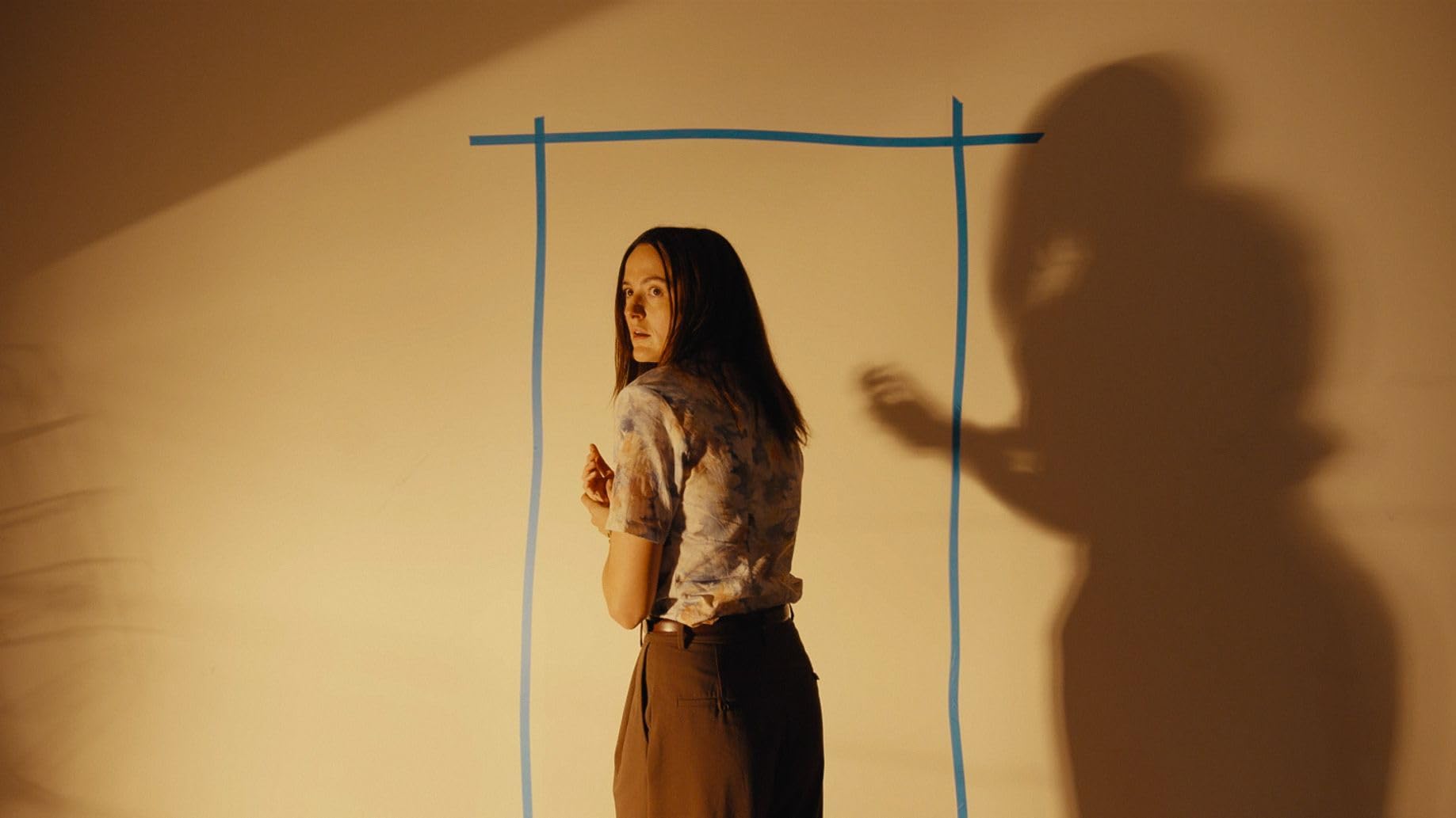 Woman in a tie-dye shirt stands inside a blue-framed rectangle painted on a beige wall, looking back toward the camera as a large shadow looms to her right.