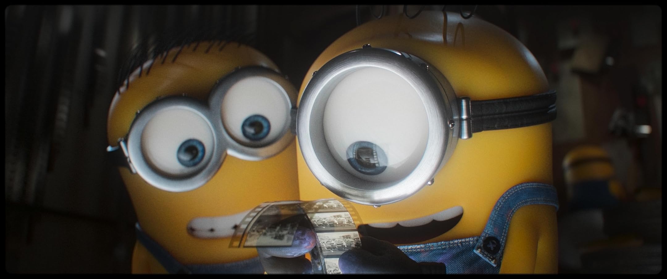 Close-up of a yellow Minion wearing large silver goggles, with denim overalls, holding a strip of film in front of its mouth.