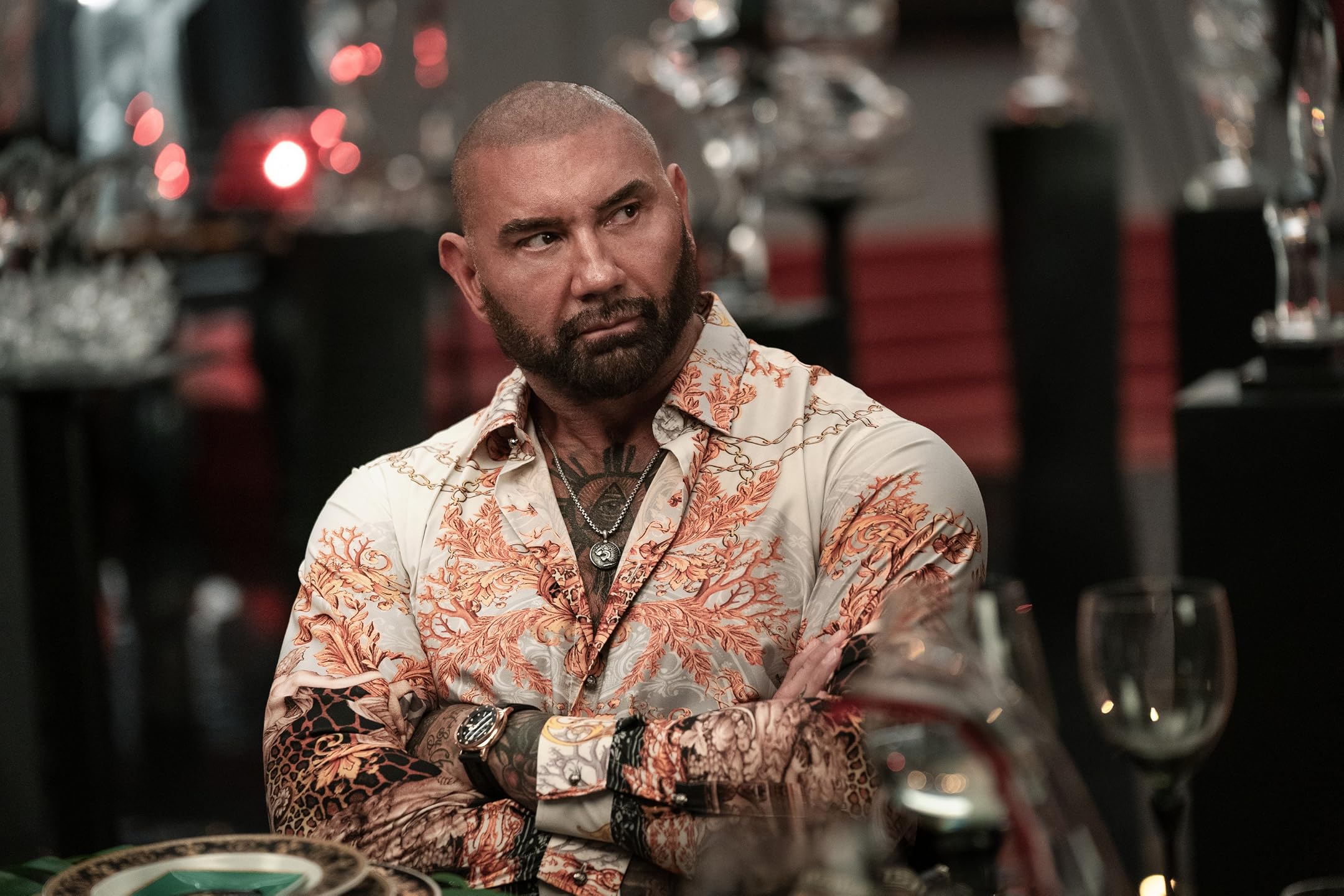Bearded man with tattoos and a coral-pattern shirt sits at a dining table in a dim, stylish restaurant setting, arms crossed.