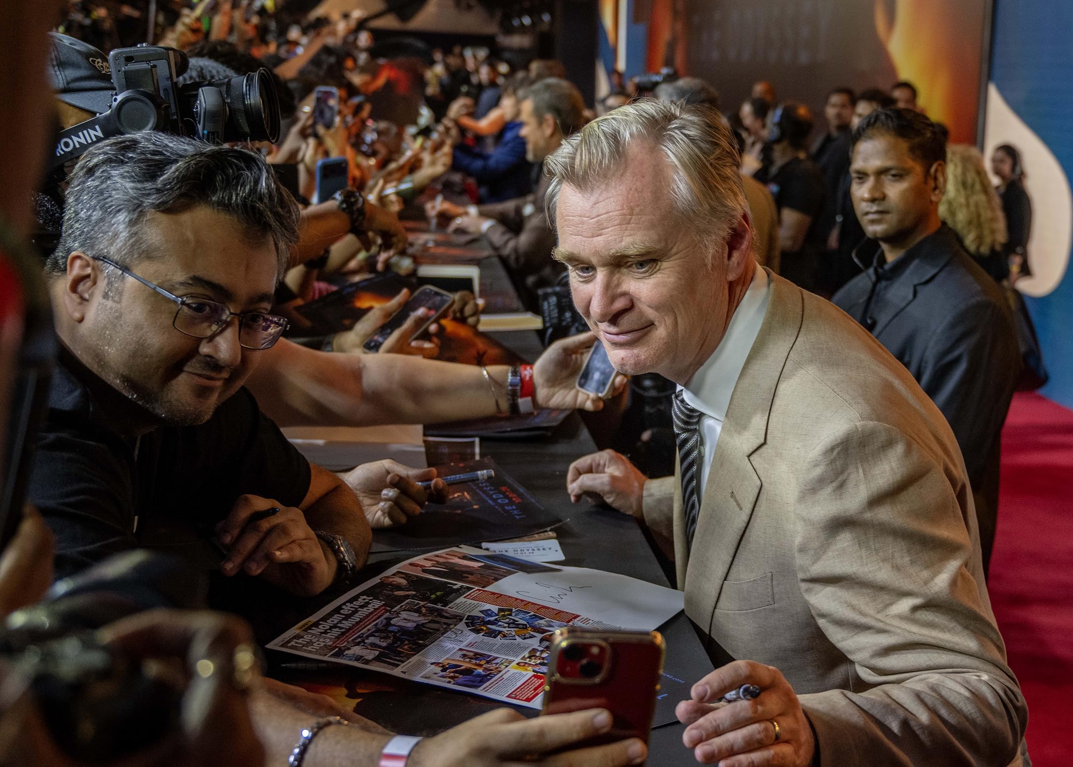 Man in beige suit signing autographs at a crowded fan event, fans and photographers in the background holding up phones and papers