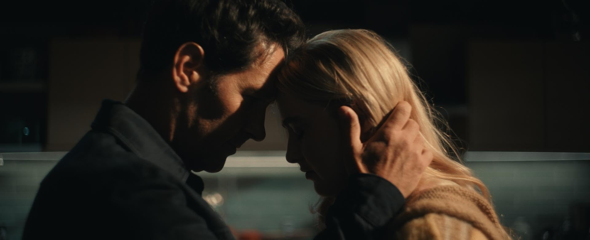 Couple leaning foreheads together in a dimly lit indoor setting, conveying an intimate moment.