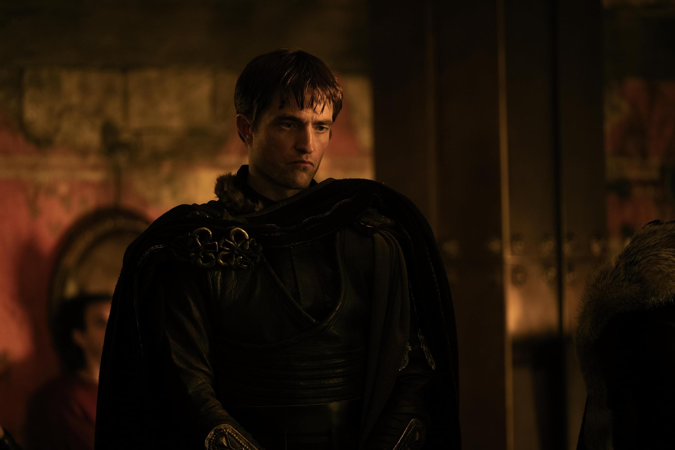 A man in dark medieval armor with a fur-trimmed cape in a dim, warm-lit hall, looking down thoughtfully.