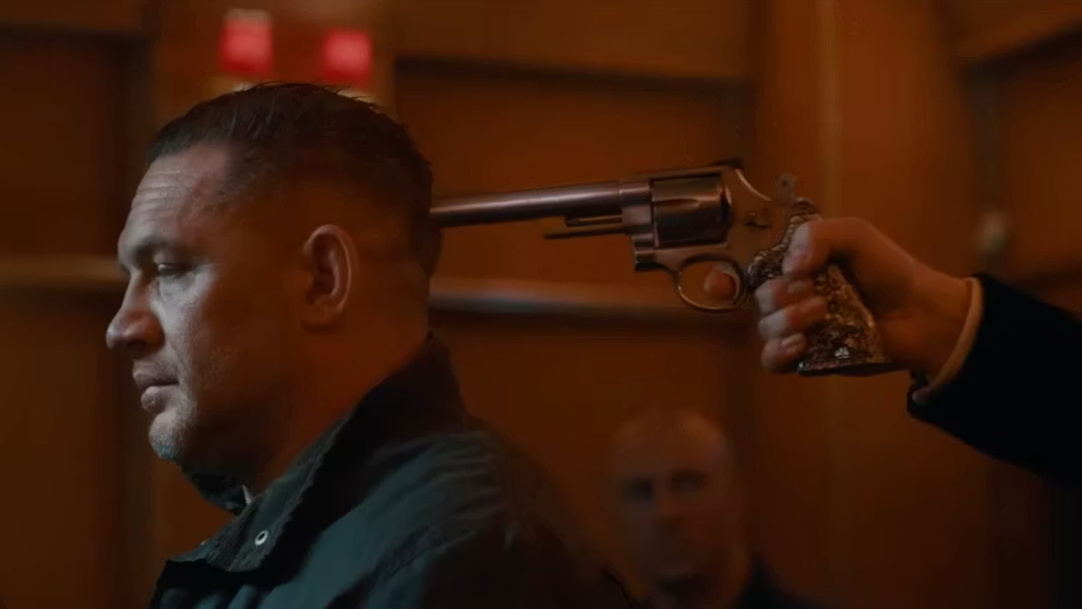 Man in profile with a revolver pressed to his temple by another person’s hand in a dim, wood-paneled room.