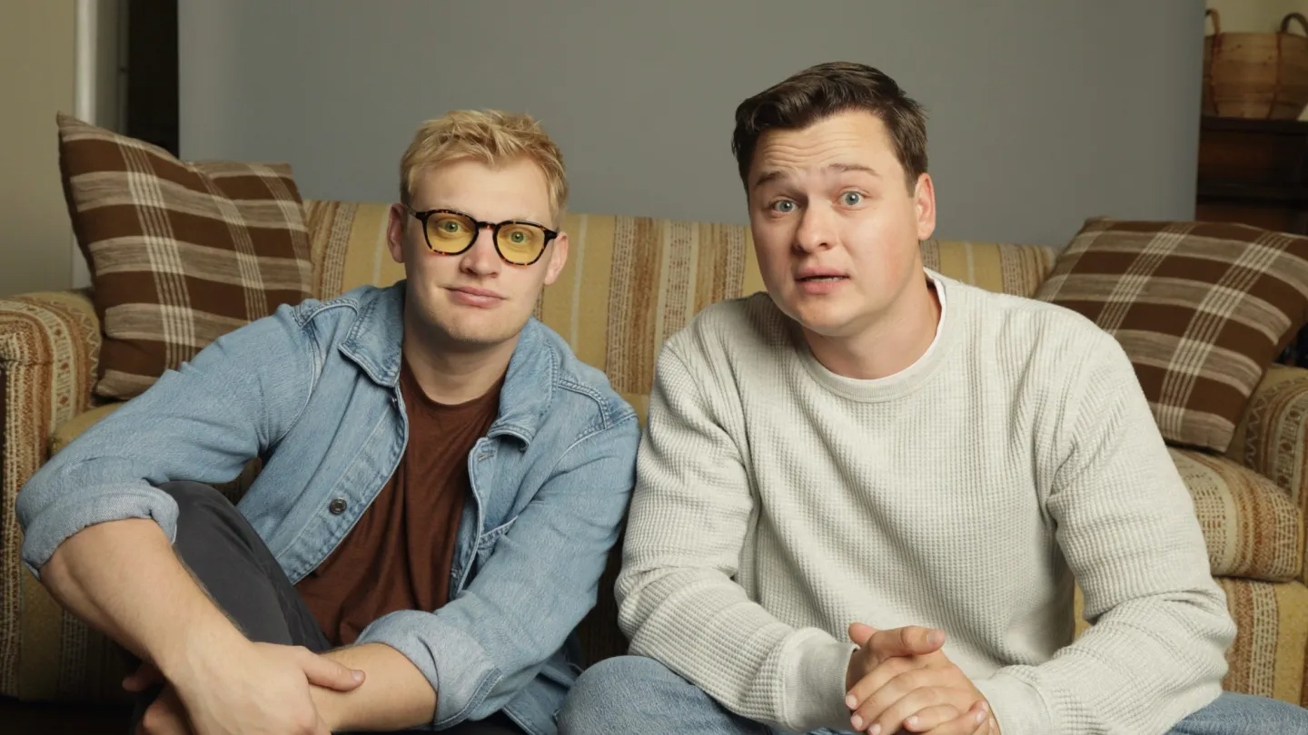 Two men sit on a striped beige couch in a living room, facing the camera; one wears a denim jacket and glasses, the other a light gray sweater.