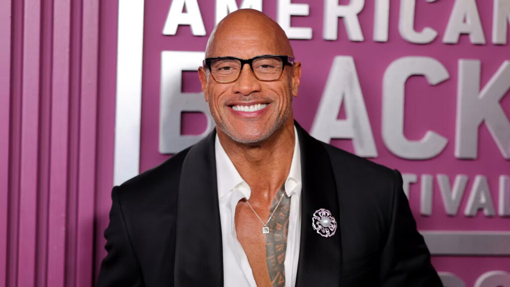 Smiling bald man in a black suit and glasses, standing before a pink backdrop with white lettering.