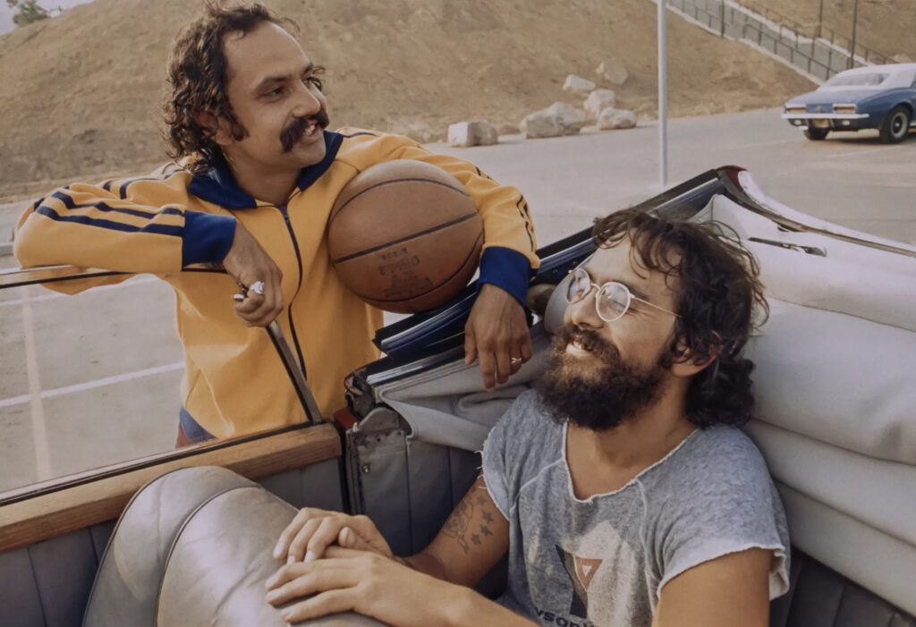 Two men posing in an open car trunk outdoors; one wearing a yellow jacket holds a basketball and smiles at the camera, the other man with a beard sits nearby.