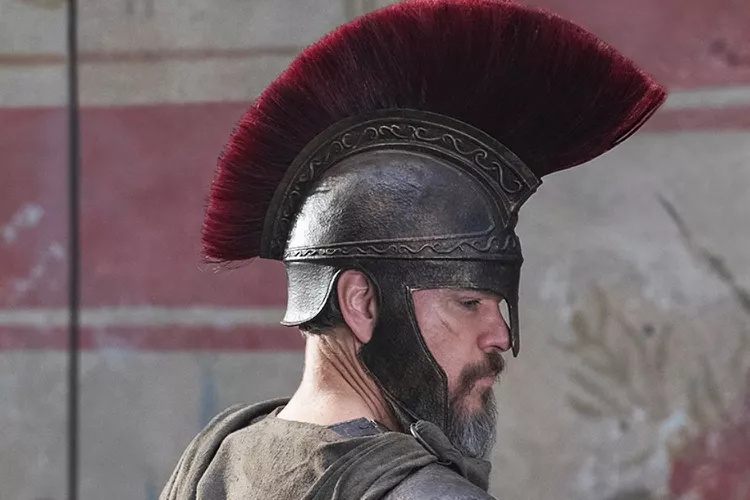 Man wearing a metal Roman-style helmet with a tall red plume, looking to the right.